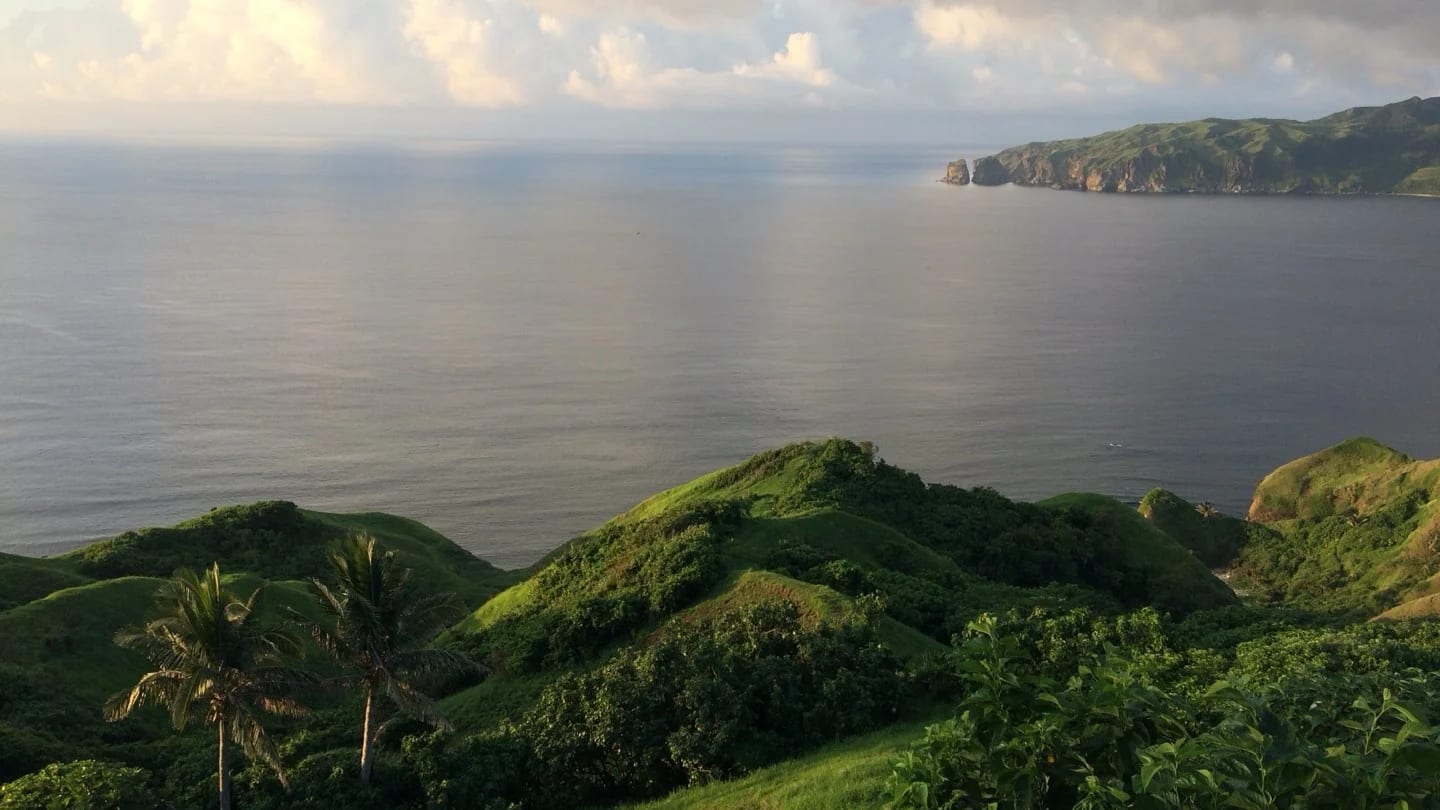 Lush green tropical hills beside an expanse of water beneath a cloudy sky.