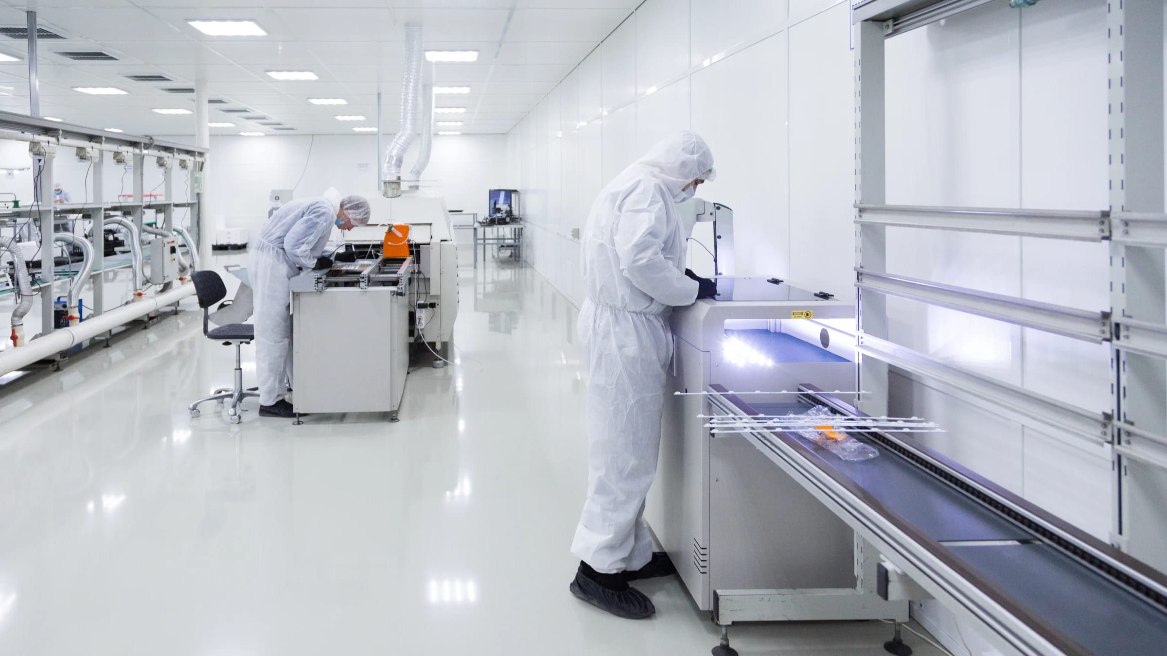 Two workers in protective clean-room clothing operate equipment inside a controlled manufacturing facility.