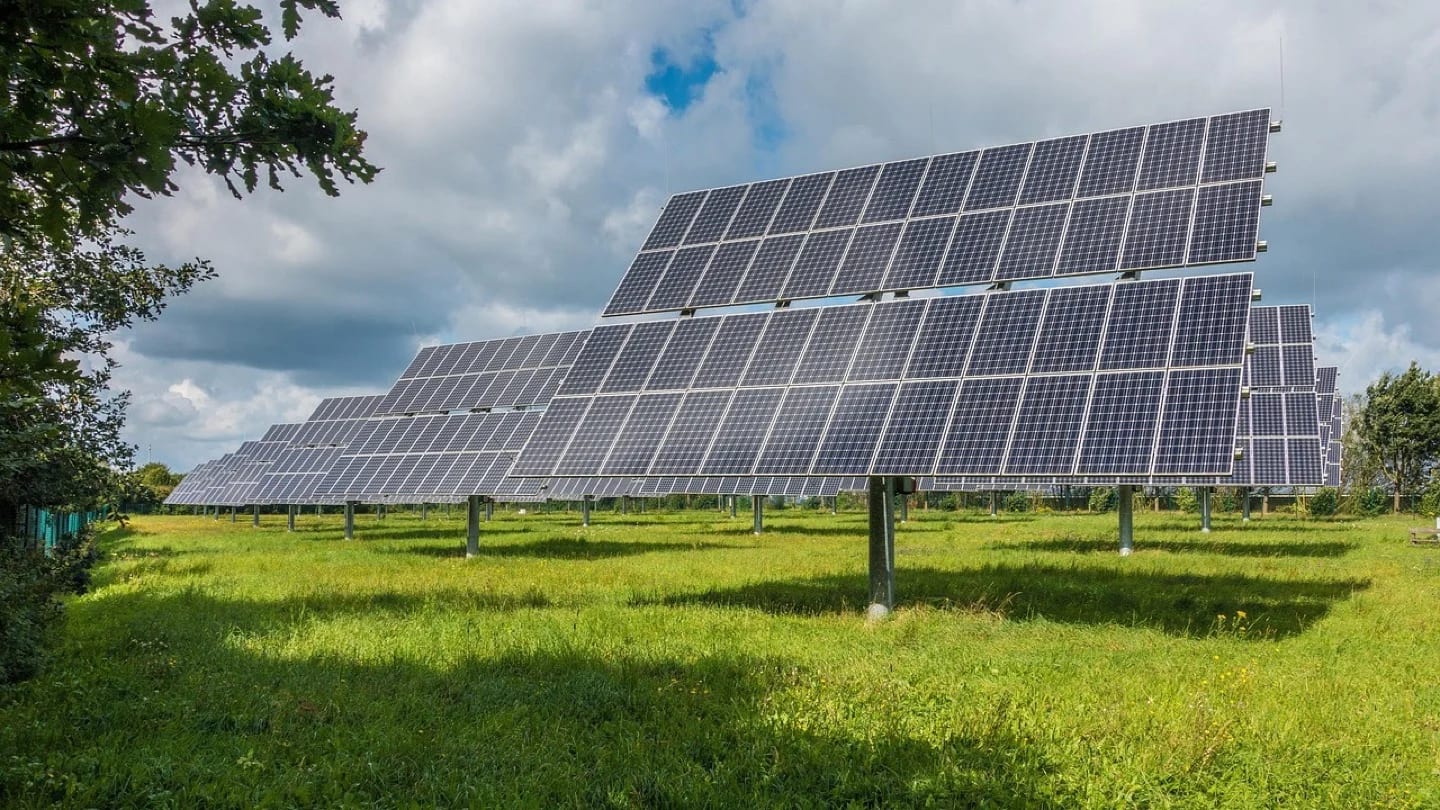 Rows of solar panels stand across a green field beneath a cloudy sky.