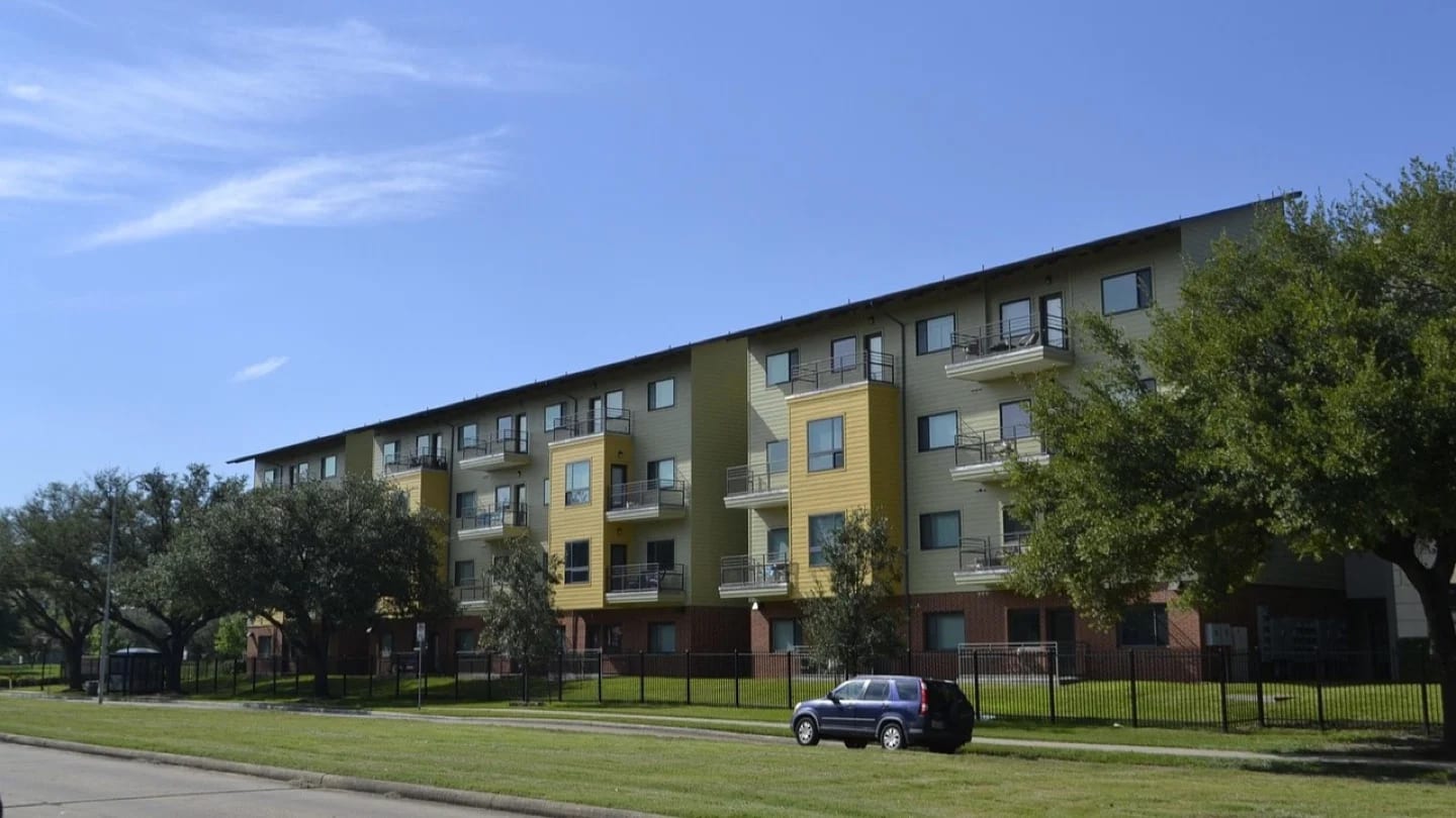 Low-rise residential apartment buildings beside grass and mature trees.