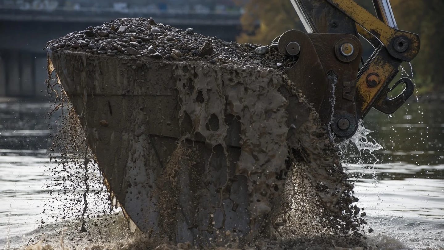 Excavator bucket lifts wet sediment and stones from shallow water.