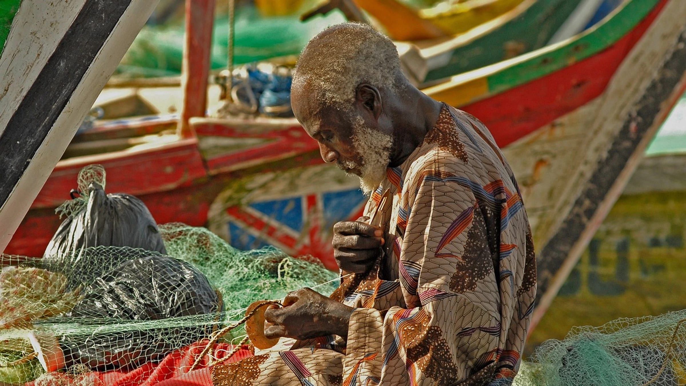 A Ghanaian fisherman repairs a net beside brightly painted fishing boats.