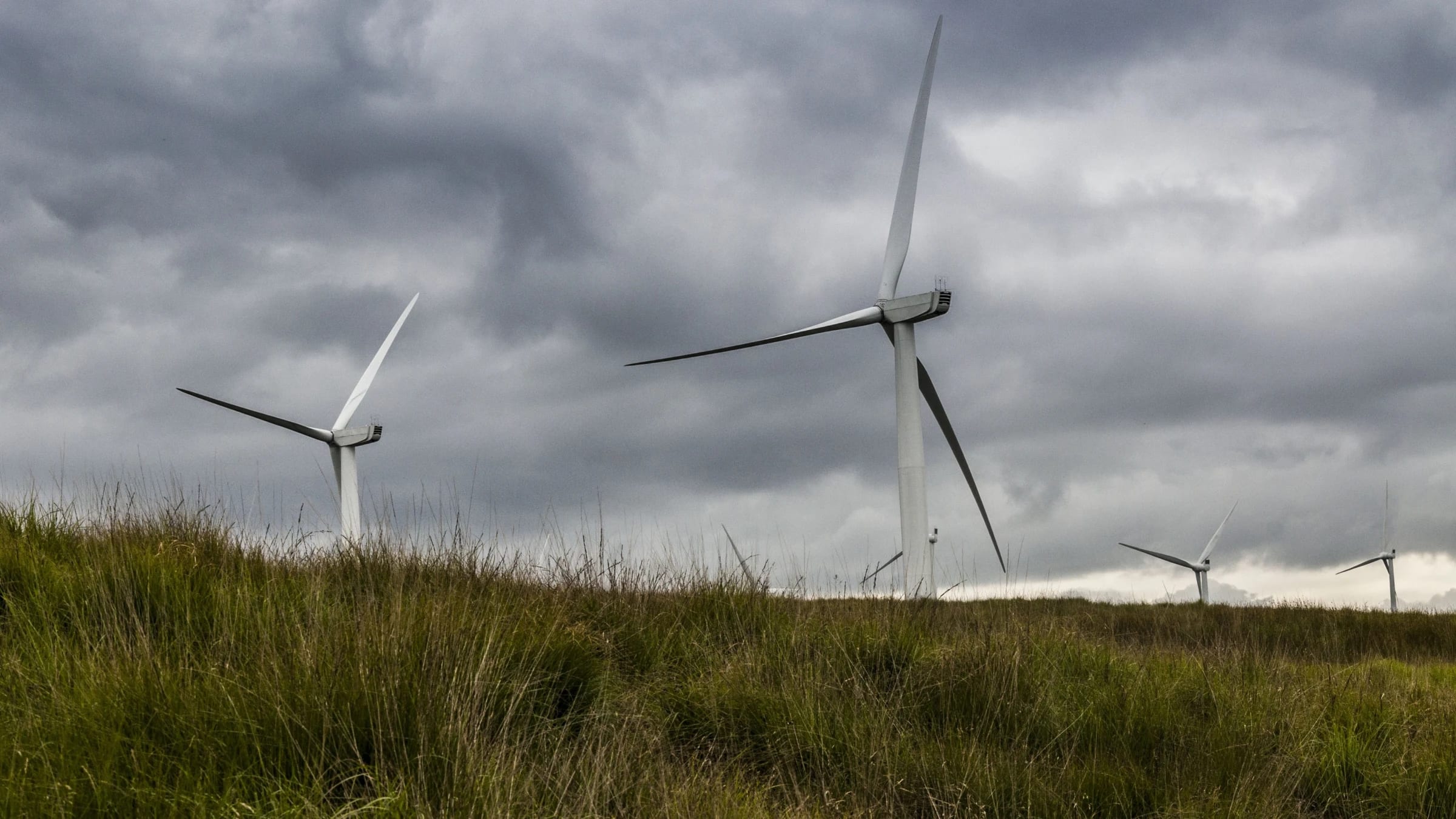 Wind turbines stand across farmland beneath a lightly clouded sky.