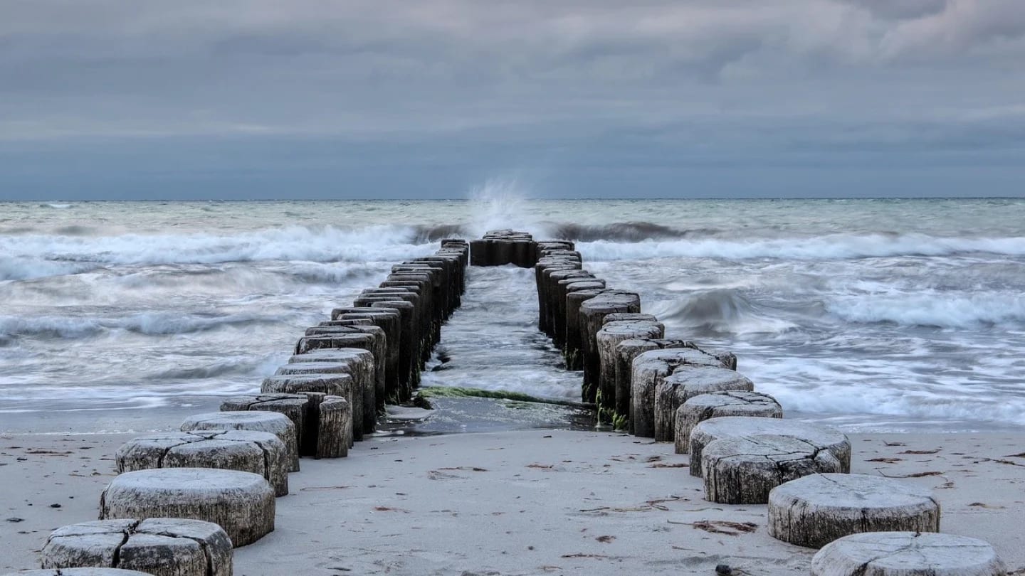 Rows of timber coastal barriers meet rough waves under a grey sky.