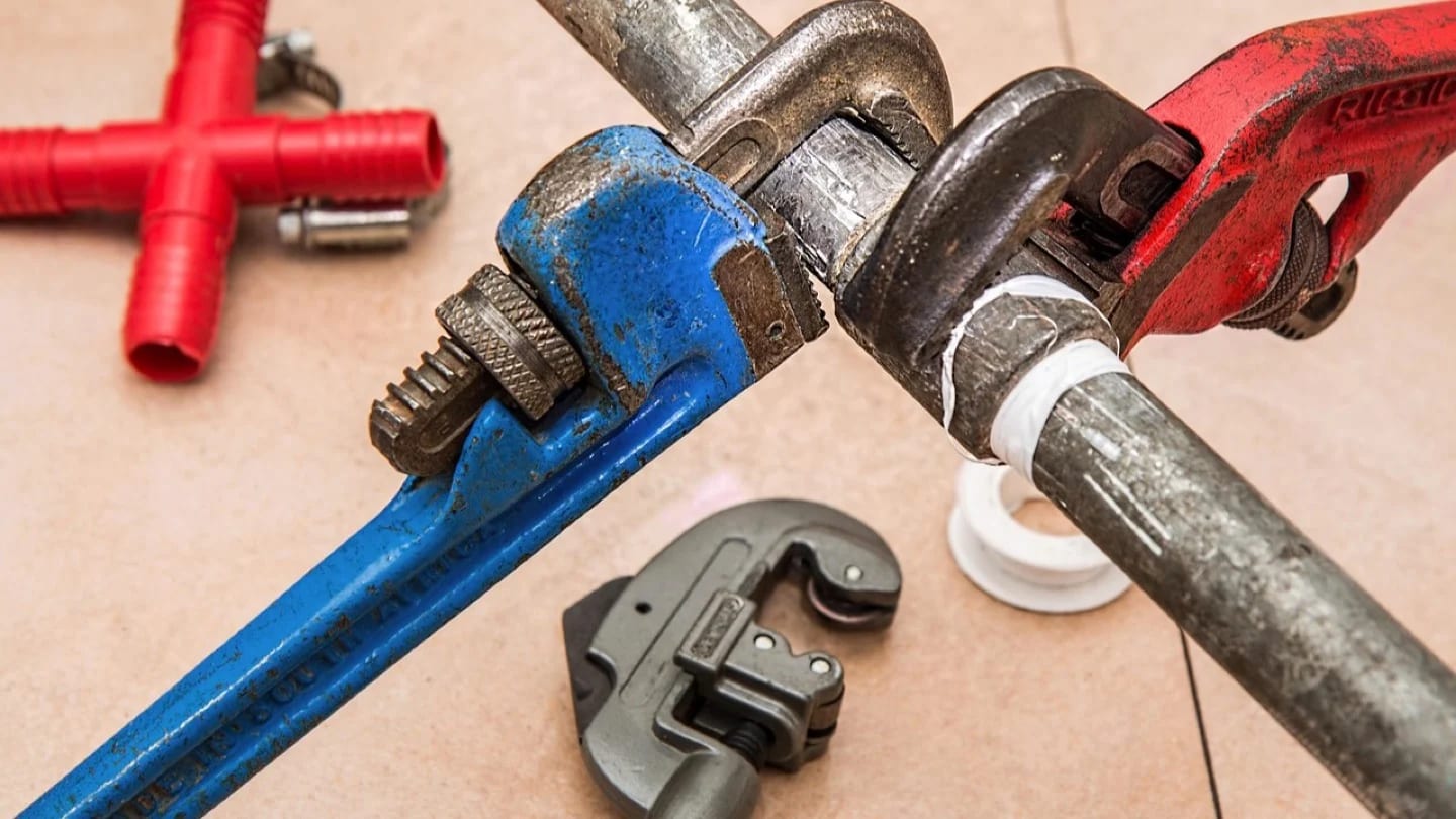 Three pipe wrenches grip a metal water pipe beside fittings and sealing tape.