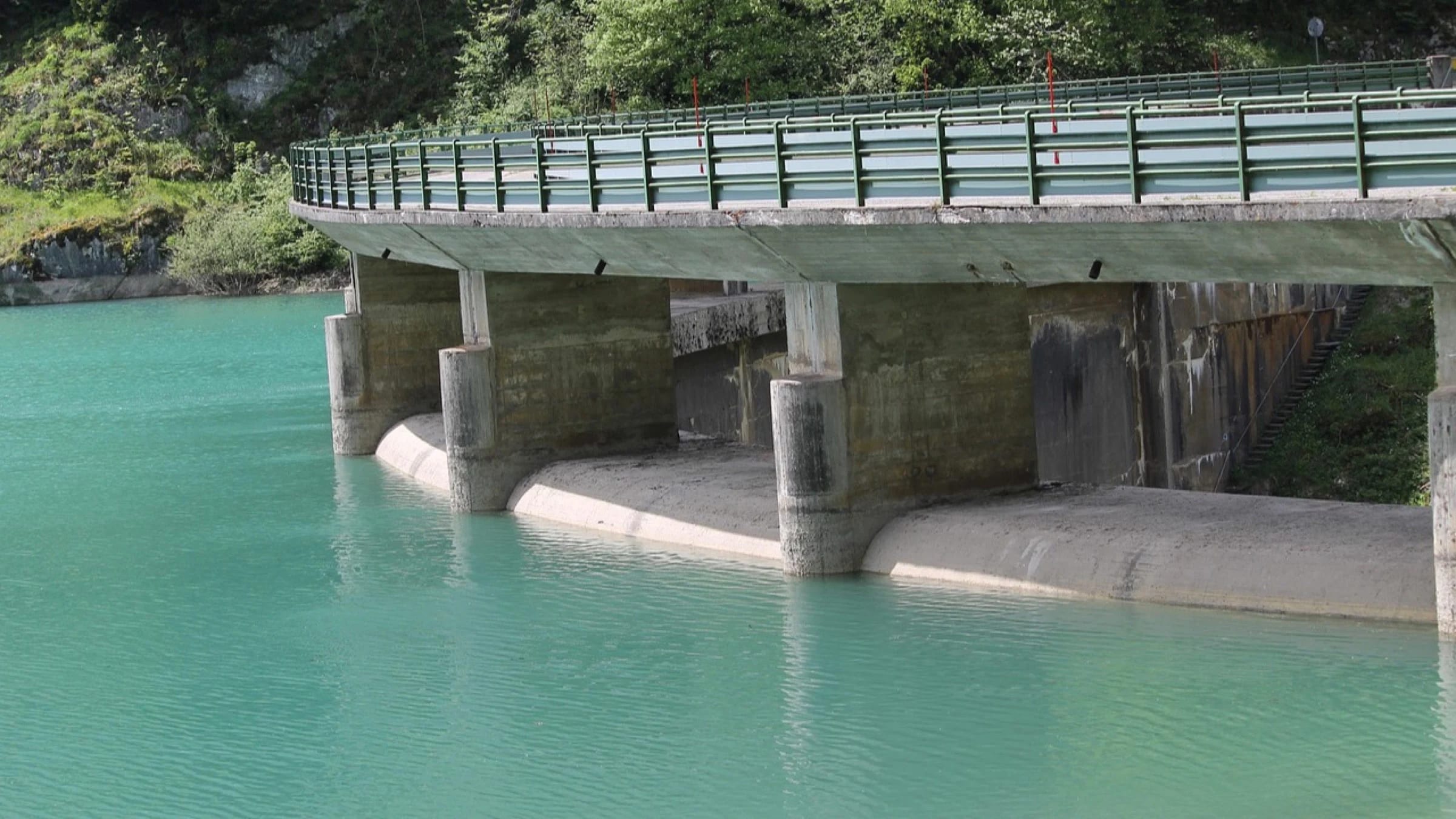 Turquoise reservoir water beside a concrete dam structure and wooded bank.