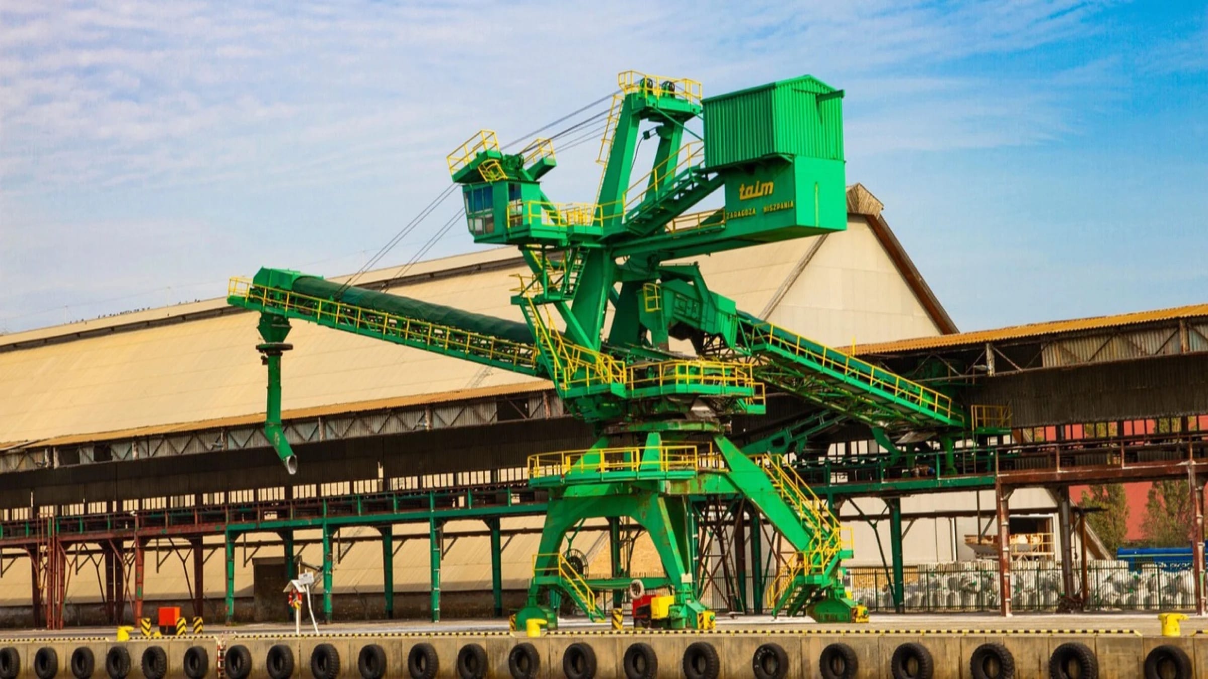 A large green dockside crane outside an industrial fabrication hall.