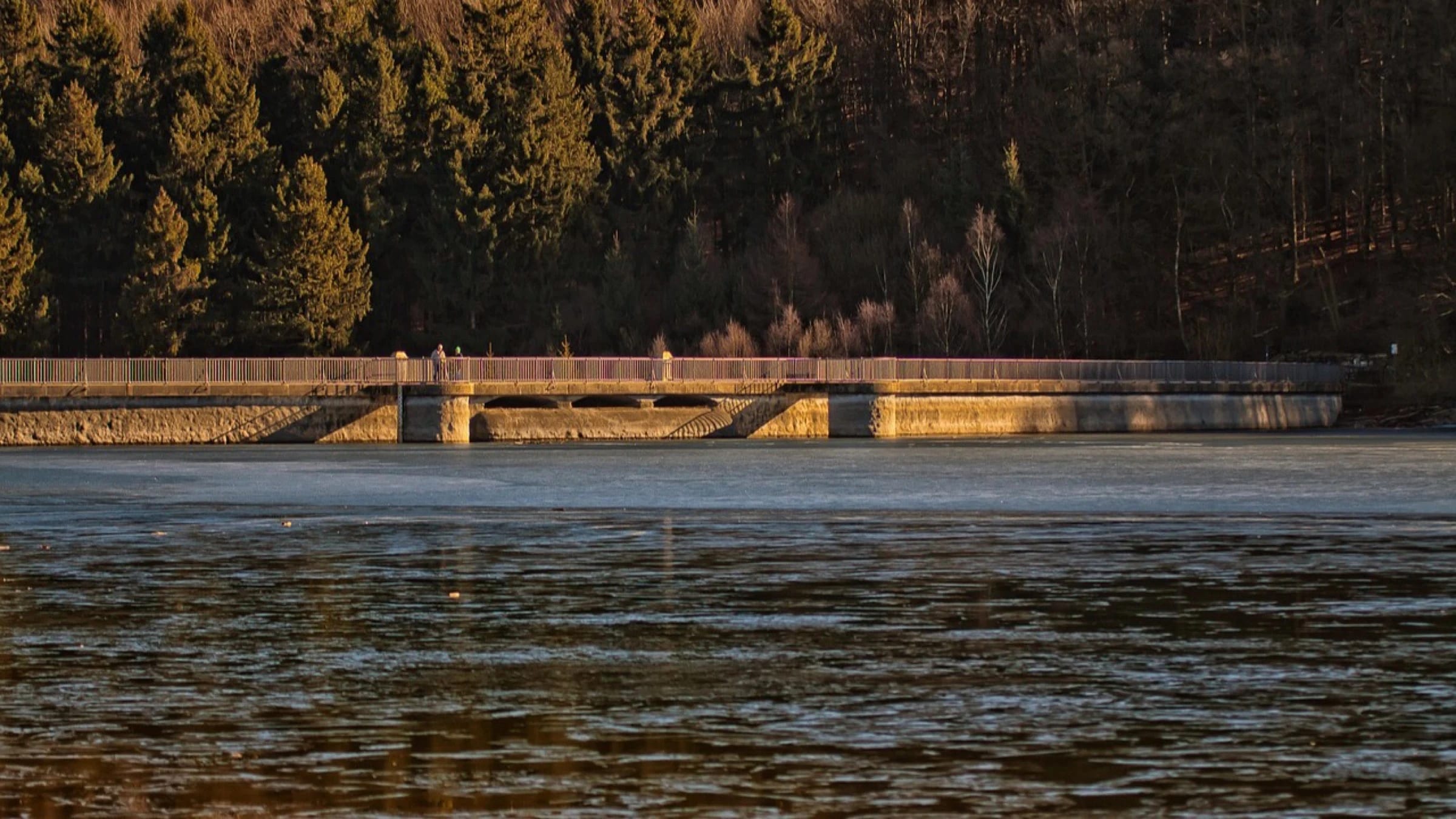 A low dam structure crossing a reservoir between wooded hills.