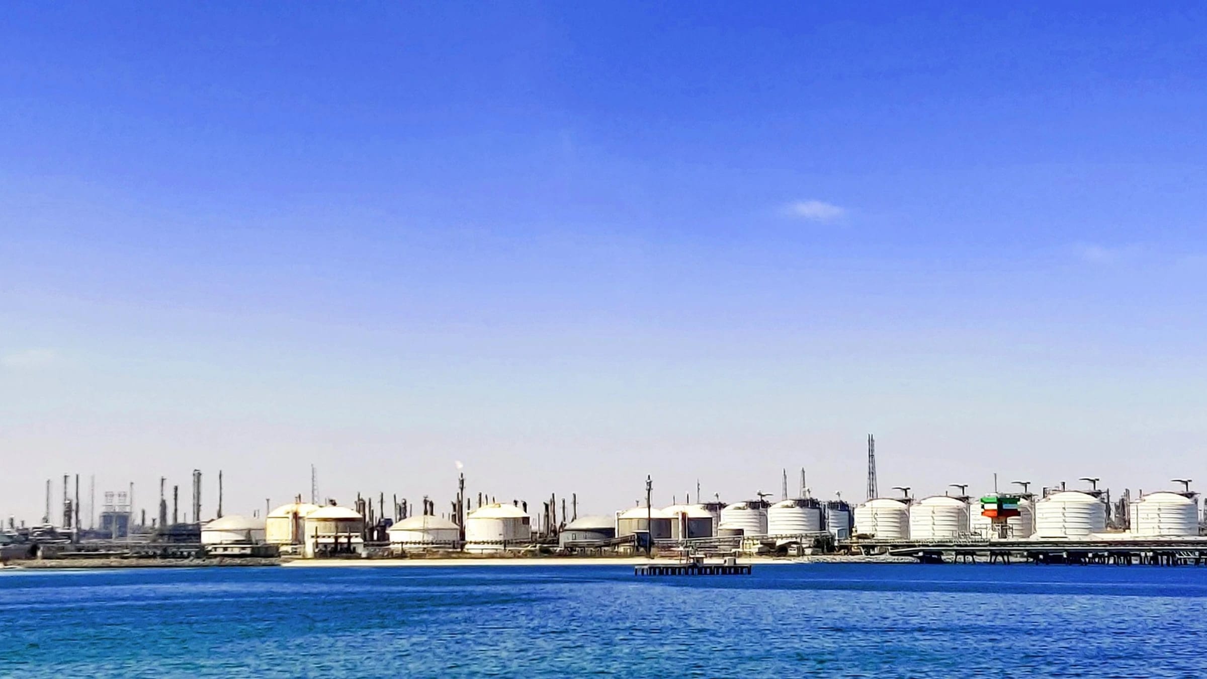 White oil-storage tanks and refinery structures beside coastal water.