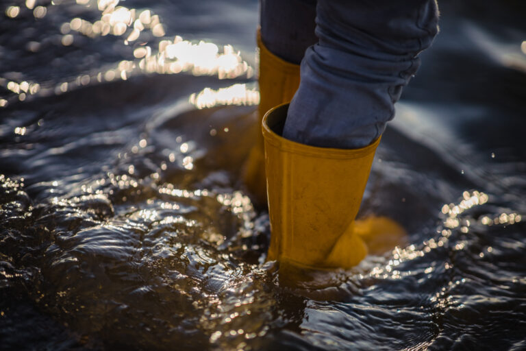 Person wearing yellow boots standing in moving floodwater, illustrating urban flooding and the need for improved flood monitoring systems.