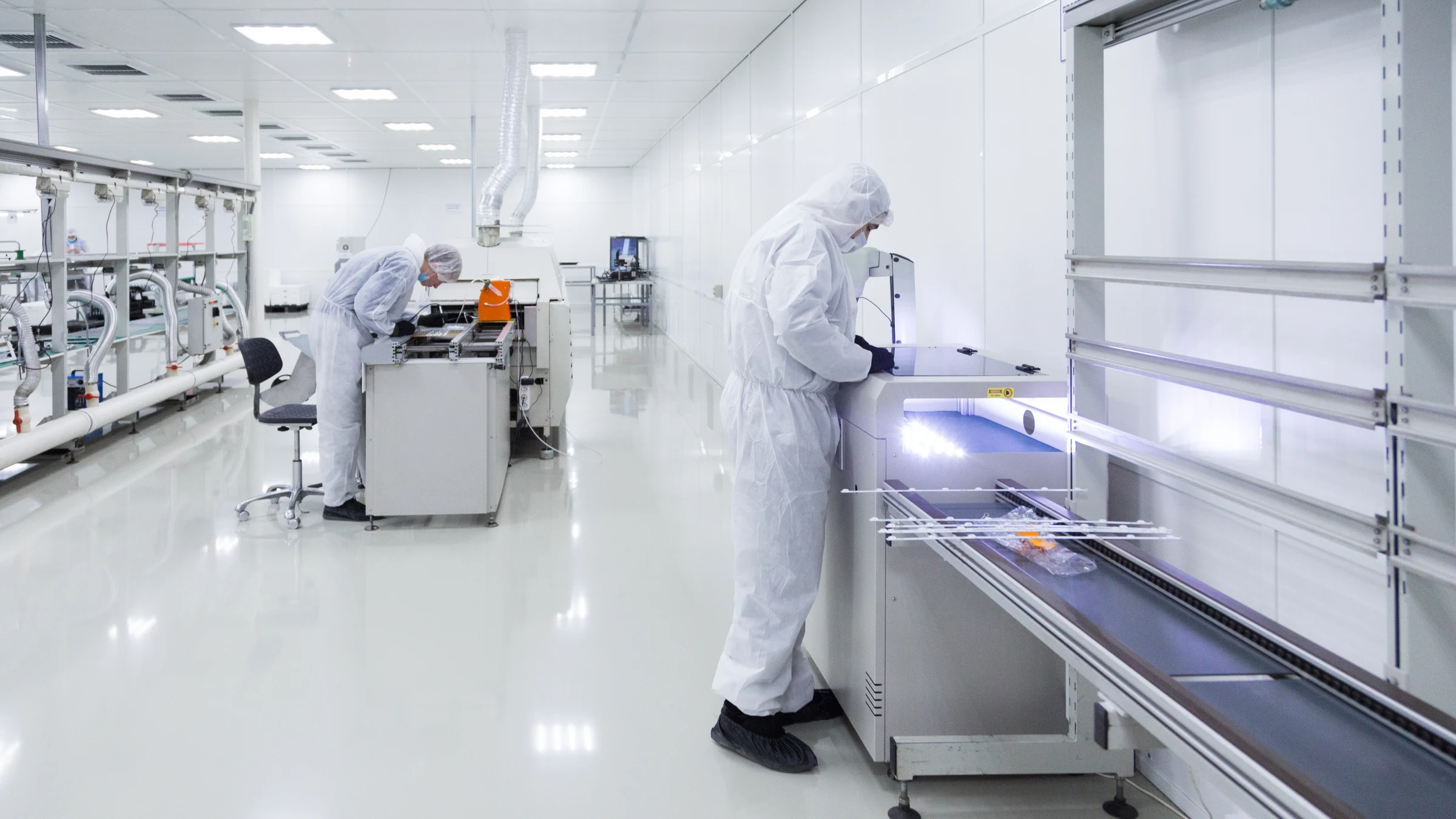 Two workers in protective clean-room clothing operate equipment inside a controlled manufacturing facility.