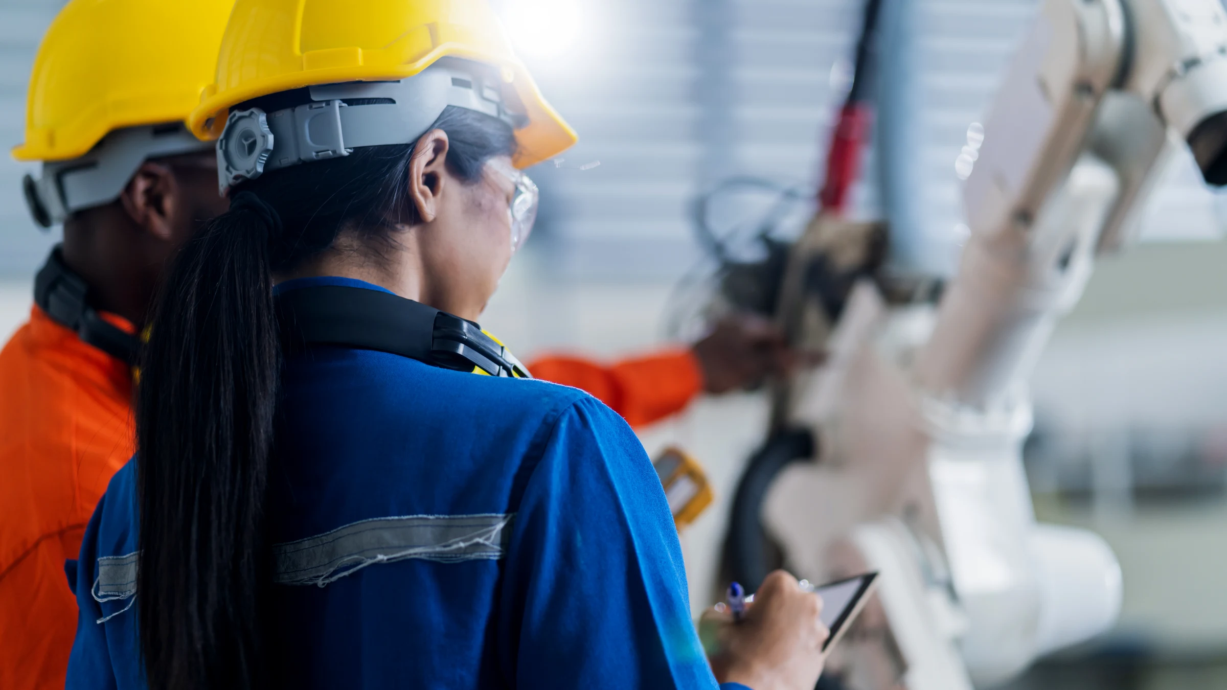 Two engineers in hard hats inspect a robotic arm inside an industrial facility.