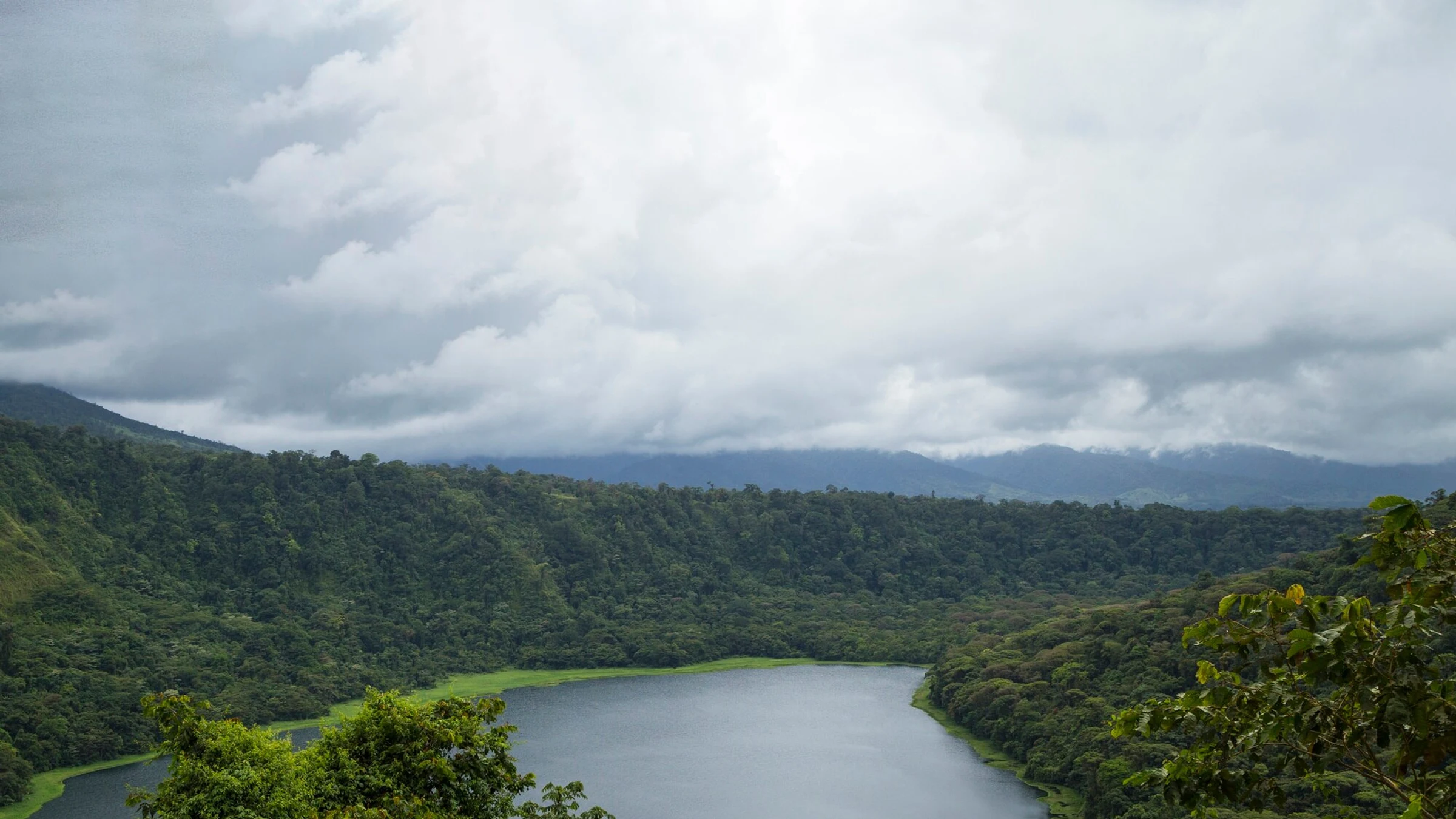 Forested hills surround a calm freshwater lake beneath a cloudy sky.