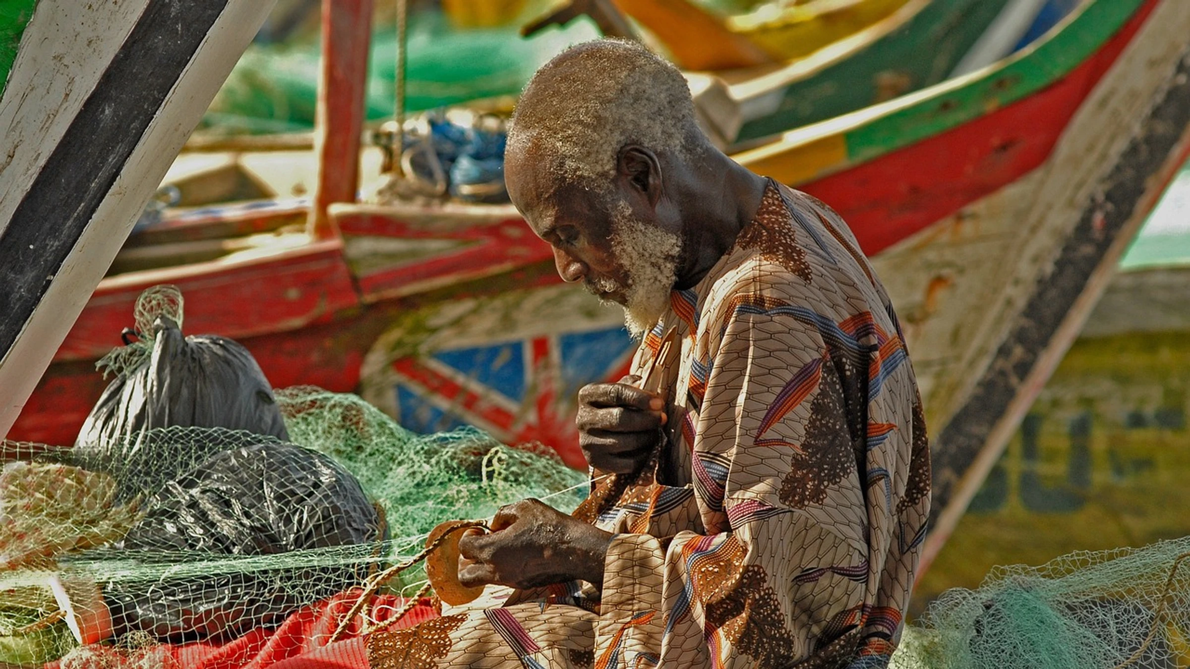 A Ghanaian fisherman repairs a net beside brightly painted fishing boats.