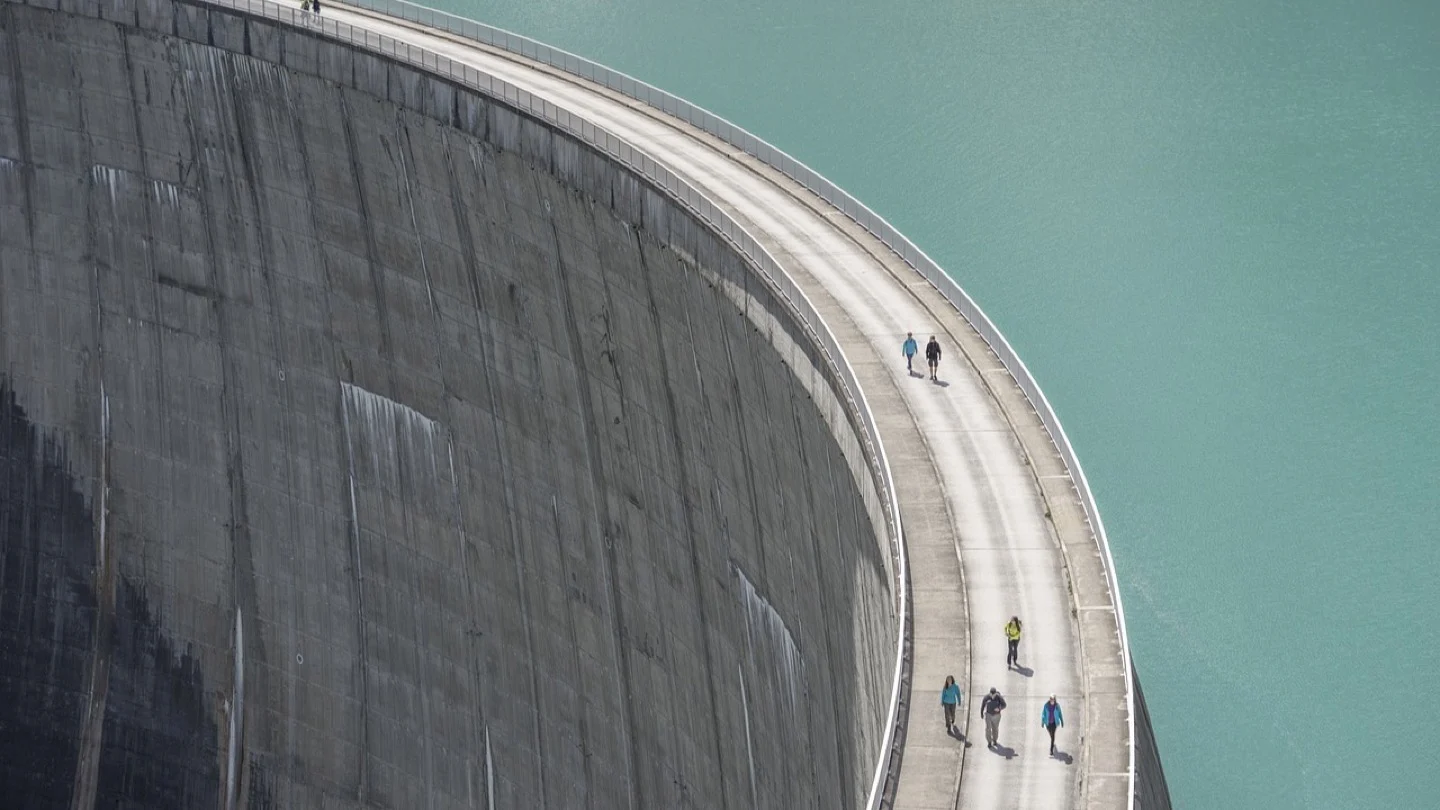 Curved concrete dam wall holding turquoise reservoir water, viewed from above.