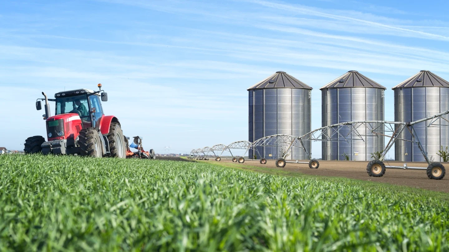 Farm machinery operates beside grain silos and irrigation equipment across cultivated fields.
