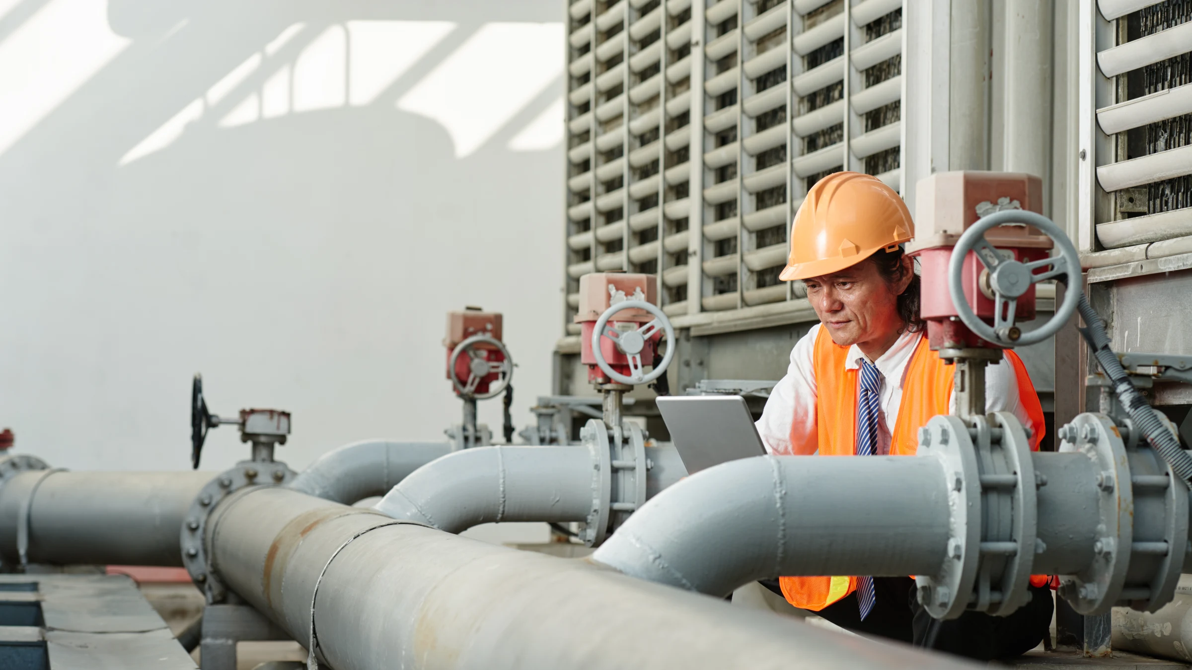 Utility worker wearing a hard hat consulting a tablet among large pipes and valves.