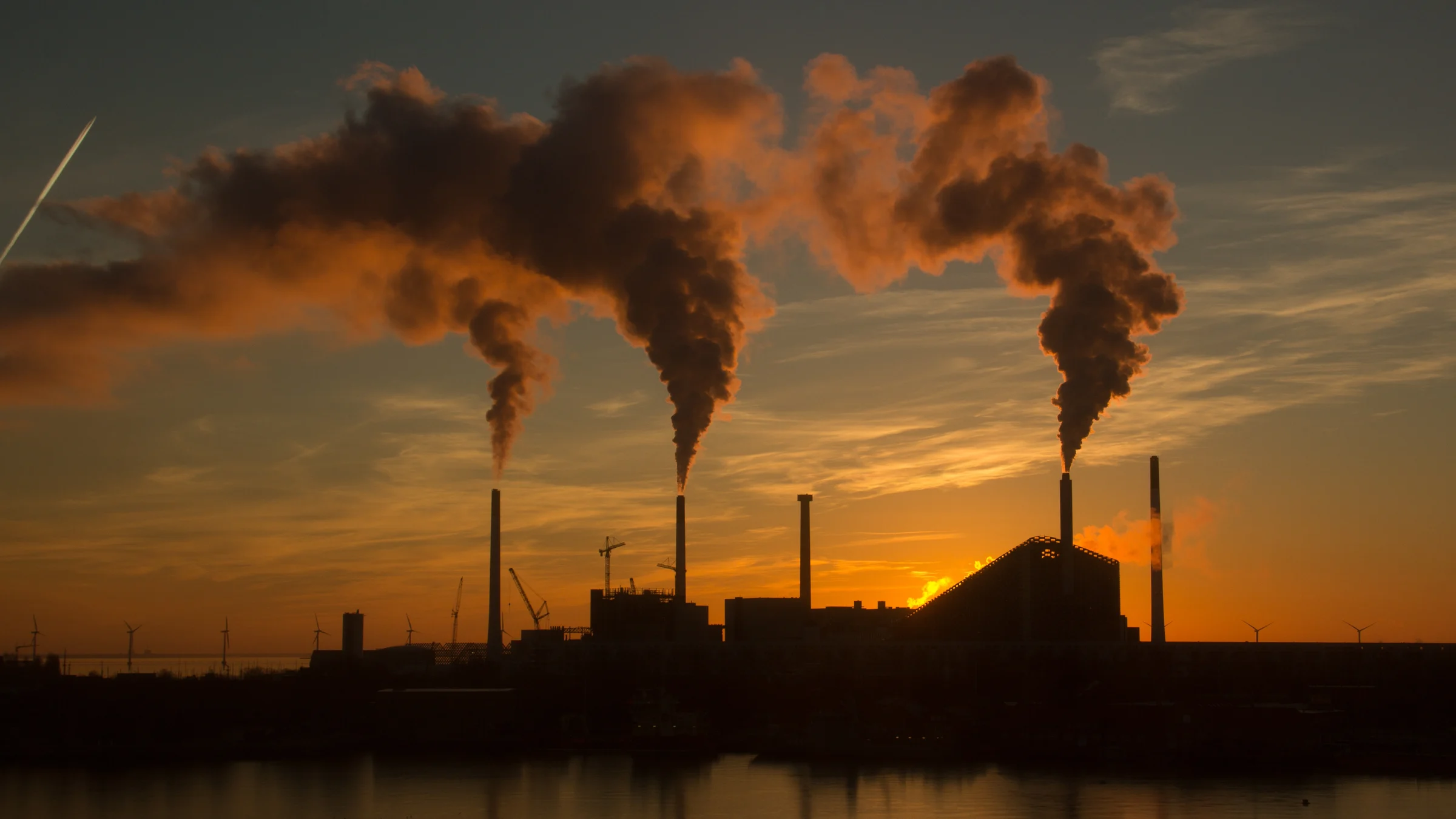 Industrial stacks and pipework release steam against a sunset sky.