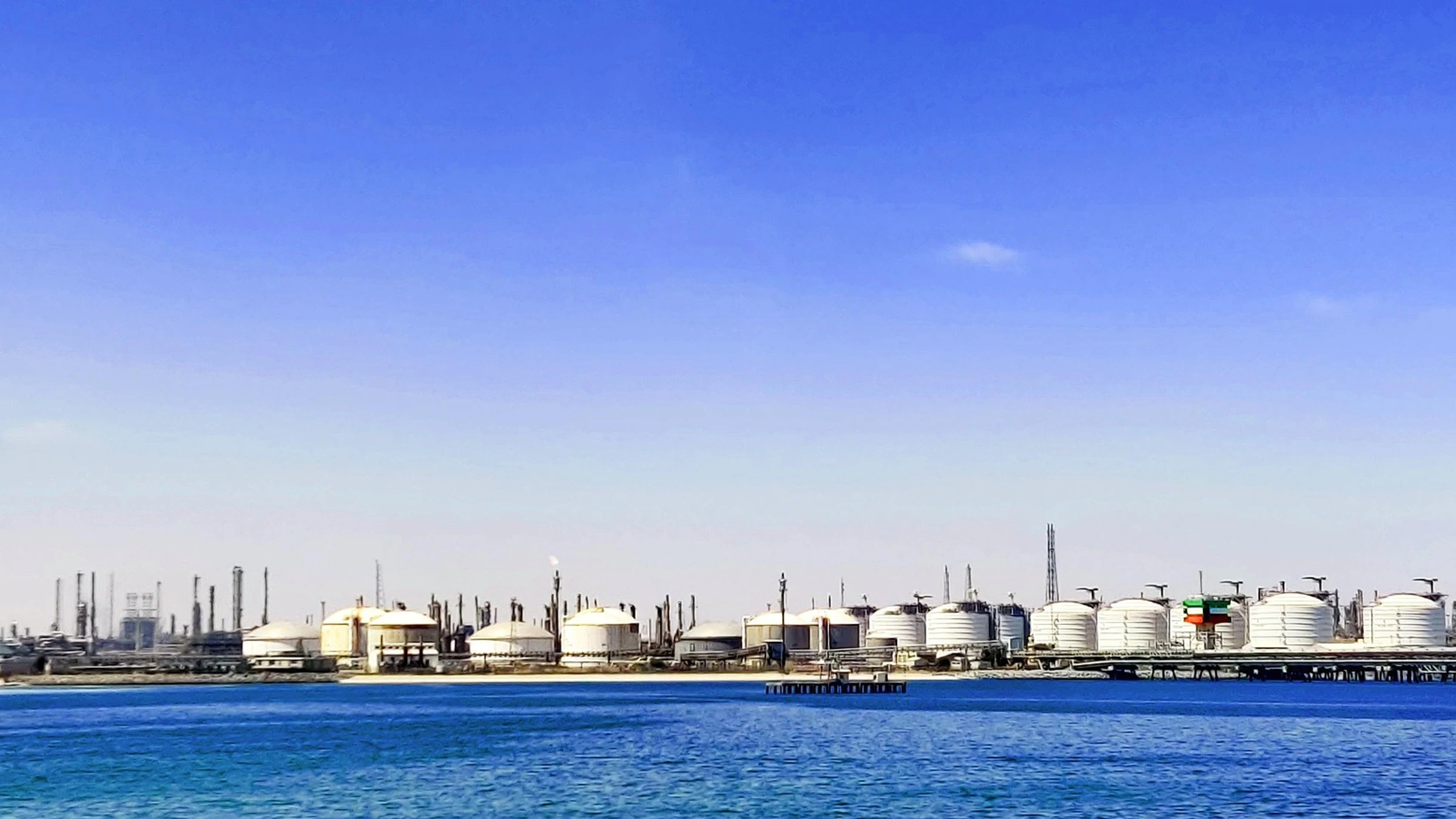 White oil-storage tanks and refinery structures beside coastal water.